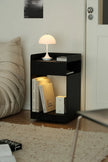 A modern black open-sided side table or nightstand in a bedroom setting, styled with a white mushroom lamp, books, and a small box. The table is positioned next to a bed with a beige pillow, on a light-colored rug, with a white door in the background.