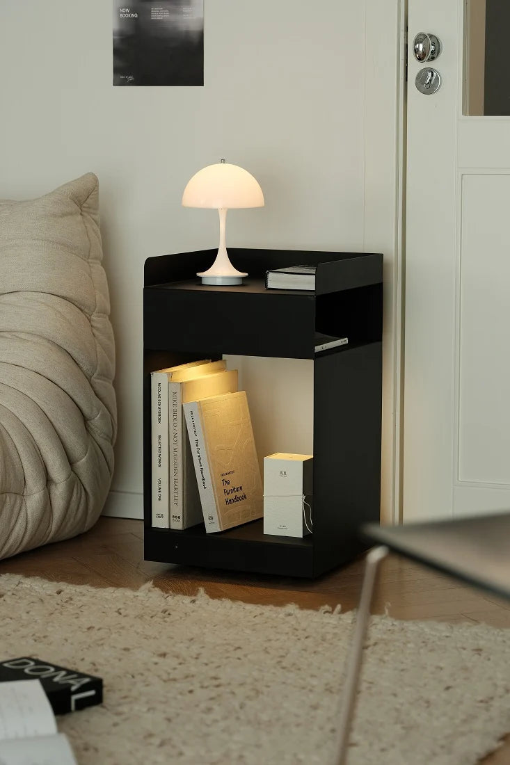 A modern black open-sided side table or nightstand in a bedroom setting, styled with a white mushroom lamp, books, and a small box. The table is positioned next to a bed with a beige pillow, on a light-colored rug, with a white door in the background.