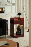 A modern dark reddish-brown wooden side table with open shelving, styled in a minimalist living room next to a black leather chair. The table is decorated with a black dome lamp, a glass of water, and books.