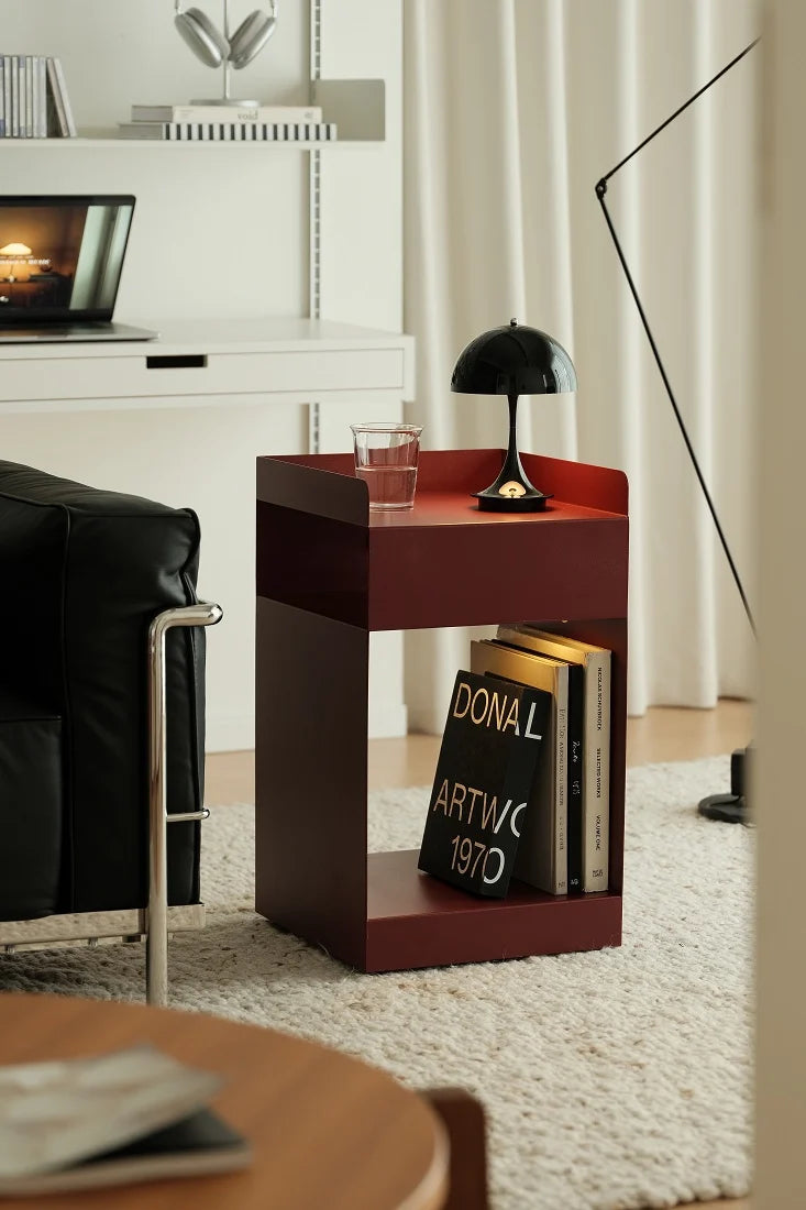A modern dark reddish-brown wooden side table with open shelving, styled in a minimalist living room next to a black leather chair. The table is decorated with a black dome lamp, a glass of water, and books.