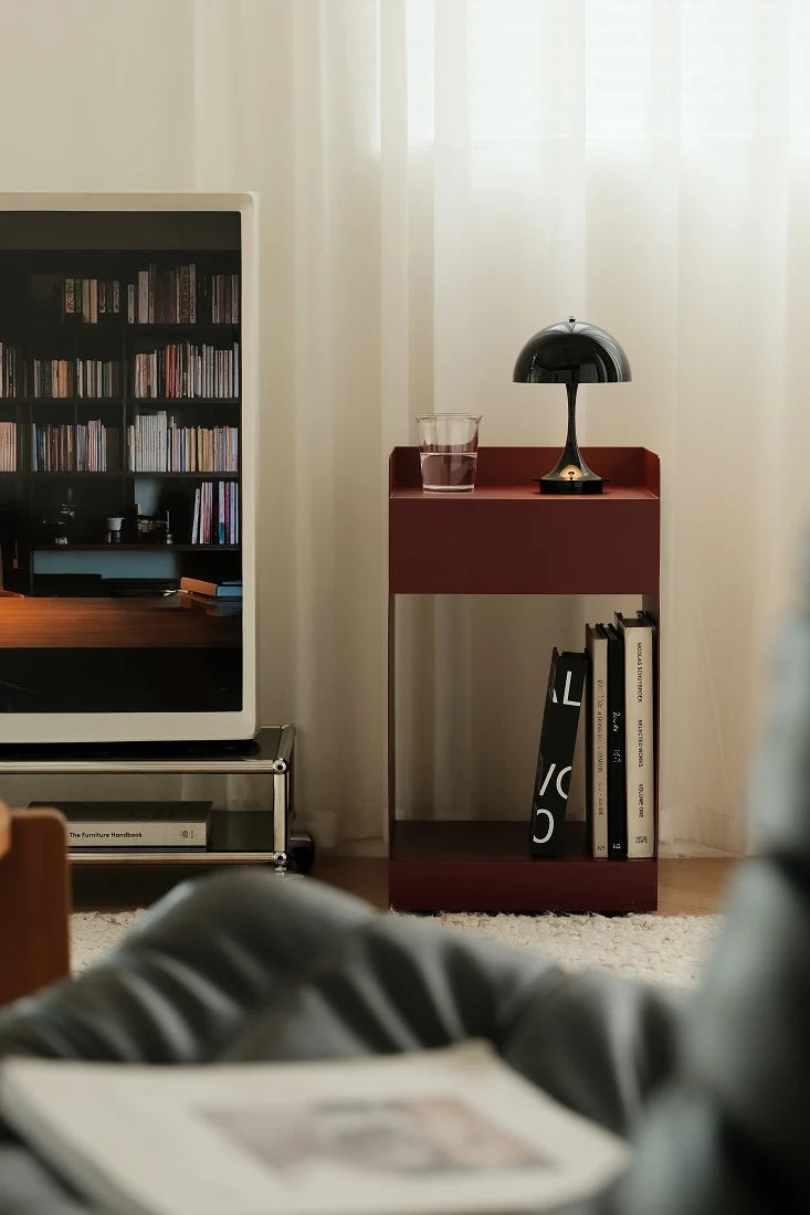 A modern dark red wooden side table with open shelving stands in a living room setting next to a television and a black dome lamp. The table is styled with a glass of water and books, set against a background of sheer white curtains.