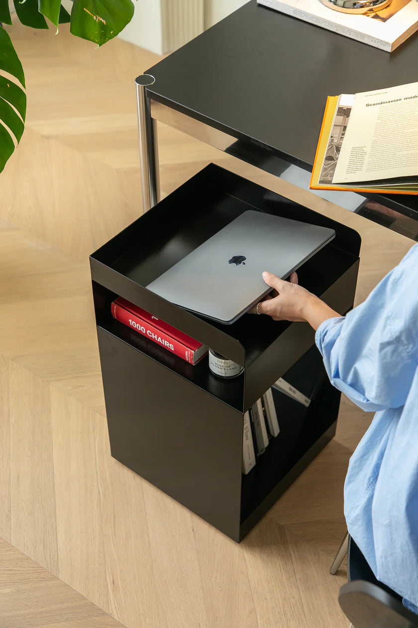 Women placing a laptop on a black small side table with books and a plant in the background.