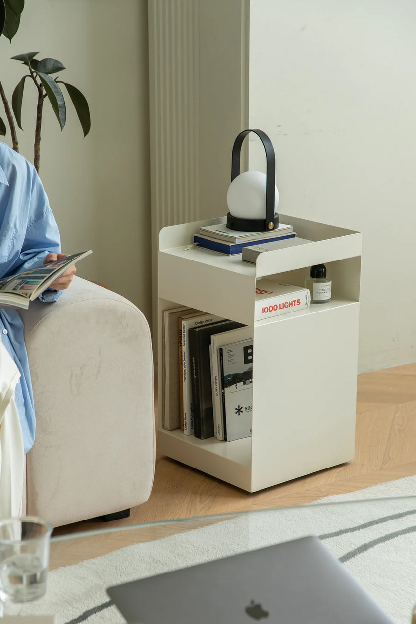 Lady sitting on a couch reading a book with a small white side table next to them holding books and a lamp.