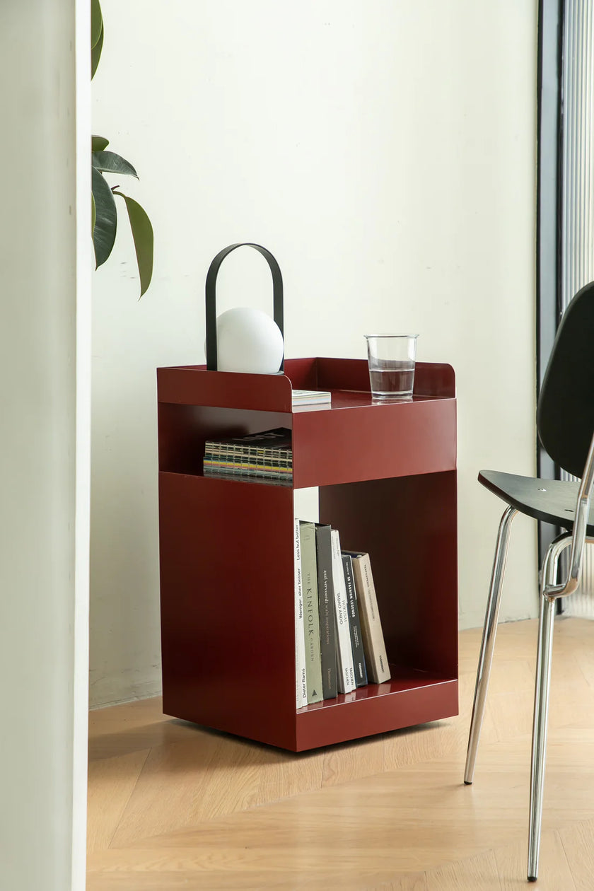 Red small side table with books, a glass, and a white object in a room with a plant and chair.