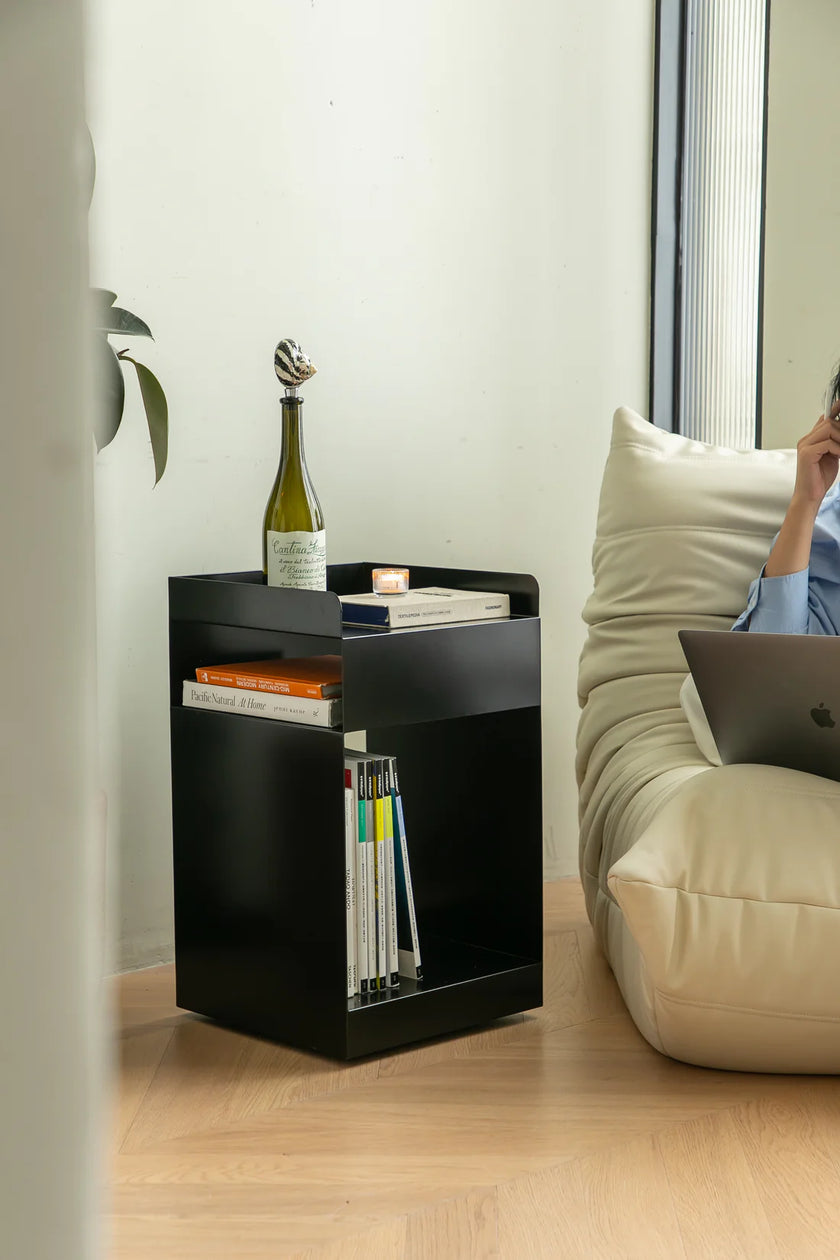 Lady sitting on a couch using a laptop next to a black sofa side table with books and a bottle.
