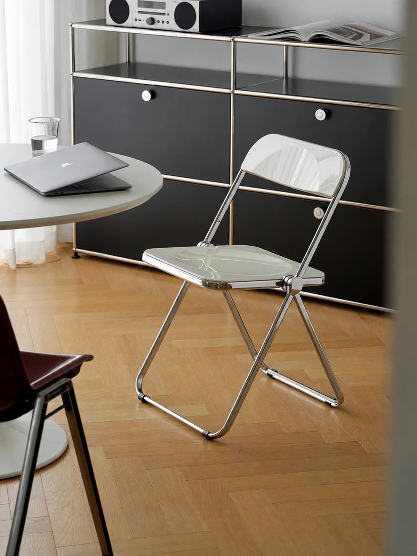 A modern chrome folding chair with a white seat and back panel, positioned on a light wood parquet floor in a contemporary room setting with a black cabinet and table in the background.