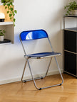 A blue folding chair with a chrome metal frame, positioned on a light wood parquet floor against a white wall in a modern room setting.