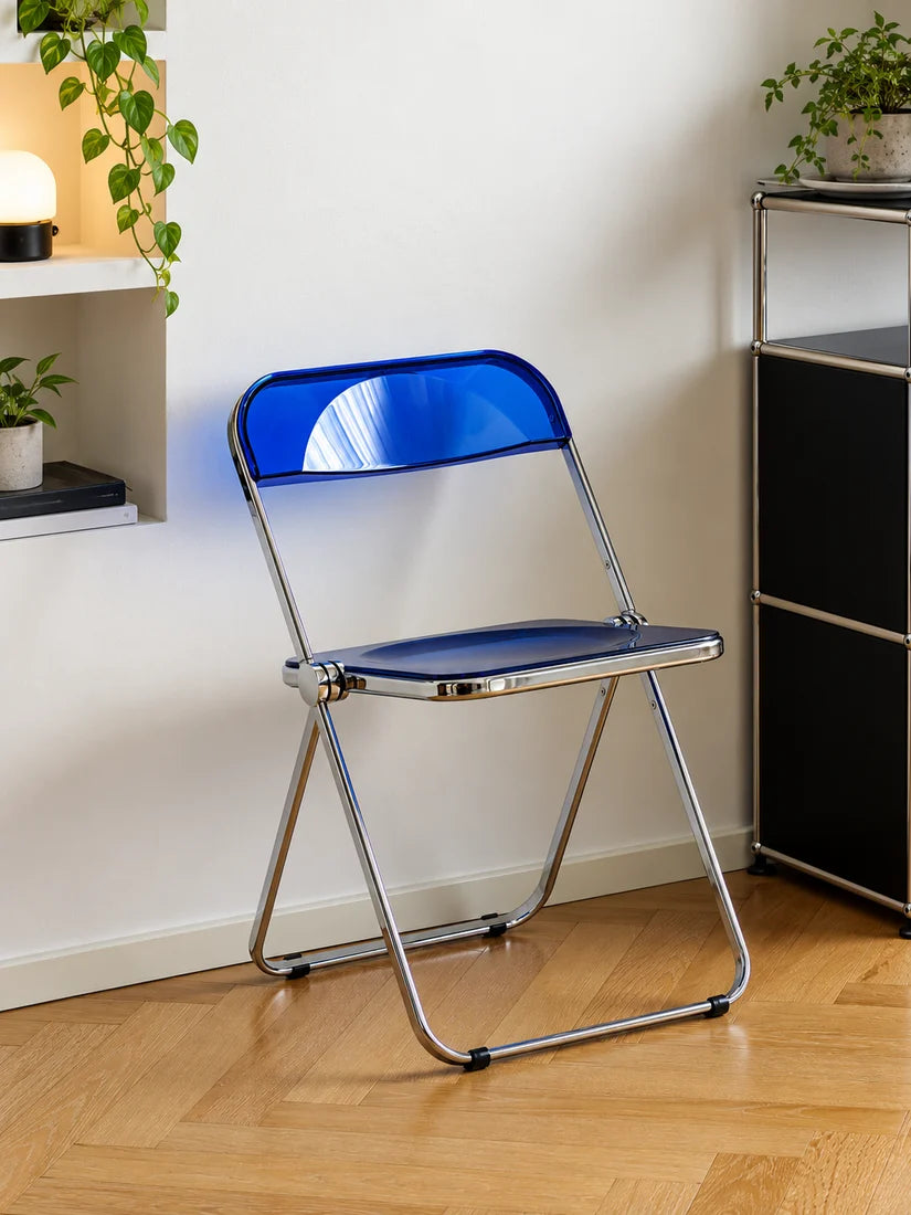 A blue folding chair with a chrome metal frame, positioned on a light wood parquet floor against a white wall in a modern room setting.