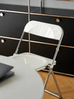 A white folding chair with a chrome metal frame, positioned on a light wood parquet floor against a white wall in a modern room setting.