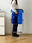 A person in a white shirt and dark pants holds a compact, folded blue chair with a silver metal frame in a minimalist room with light wood floors and a white wall.
