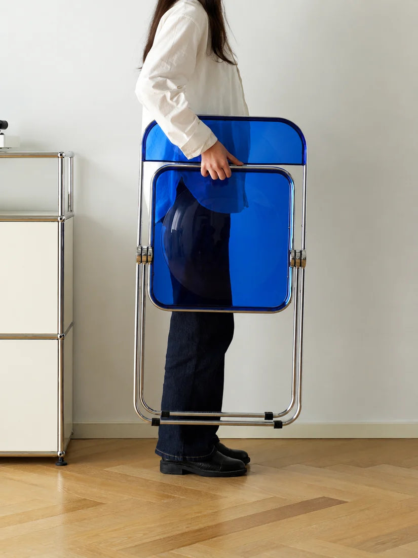 A person in a white shirt and dark pants holds a compact, folded blue chair with a silver metal frame in a minimalist room with light wood floors and a white wall.