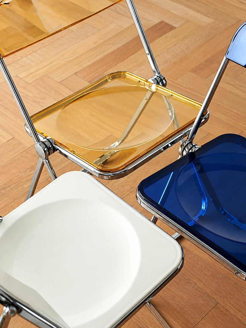 Three modern folding chairs with translucent acrylic seats in white, amber, and blue, all with chrome metal frames, arranged on a light wood parquet floor.
