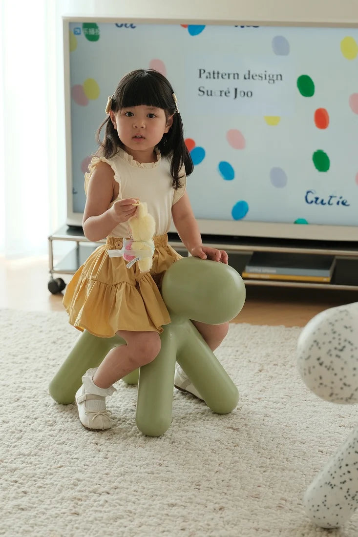 A young girl with dark hair sits on a light green octopus-shaped children's stool in a bright room, wearing a cream top and a yellow tiered skirt. A large screen in the background displays colorful polka dots and the text 'Pattern design Su:re Joo'.