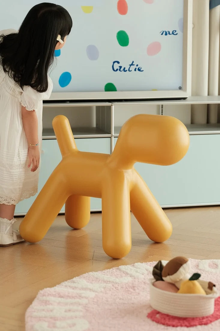 A child in a white dress stands beside a bright mustard-yellow dog-shaped stool in a modern room with a whiteboard and light blue cabinet in the background.