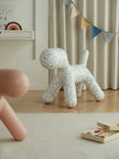 A white dog-shaped floor cushion with black speckles sits on a light wood floor in a styled children's room setting with a beige rug, curtains, and toys.