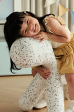 A young child wearing a yellow ruffled dress and white speckled tights hugs a matching white speckled plush toy in a brightly lit indoor room.