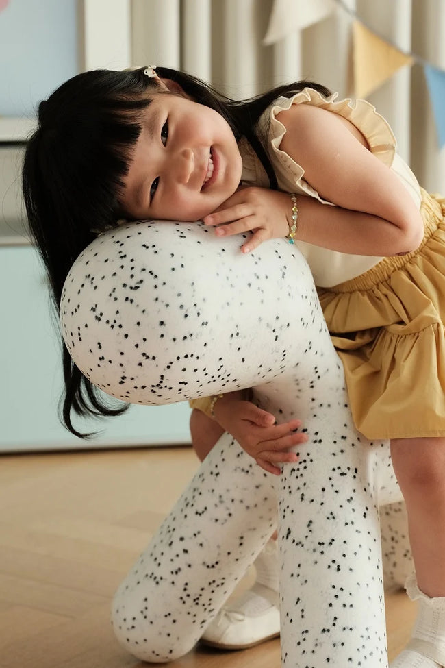 A young child wearing a yellow ruffled dress and white speckled tights hugs a matching white speckled plush toy in a brightly lit indoor room.