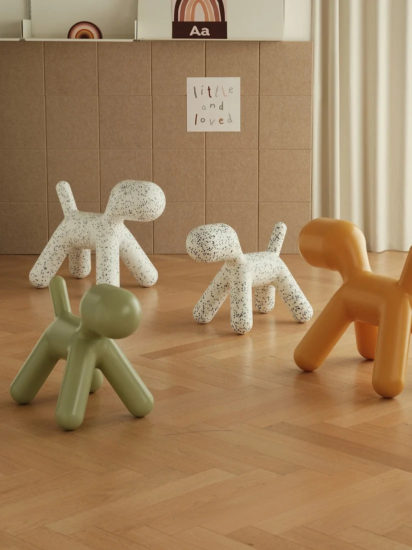 Four modern dog-shaped stools in sage green, white speckled, and mustard yellow arranged on a light wood floor in a styled interior room.