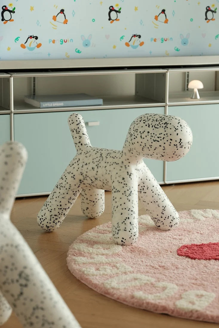 A dog-shaped white chair with black speckles, styled in a children's room with a pink rug, light wood floor, and pale blue cabinets in the background.