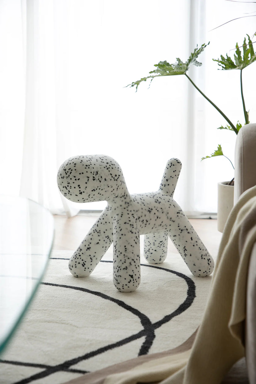 White dog-shaped plastic kids chair with black speckles on a rug in a room with plants and a chair.