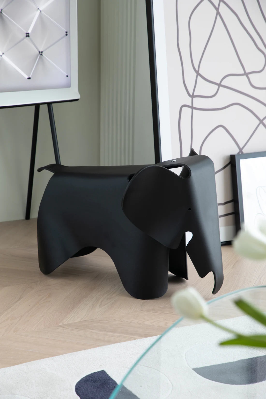 Black elephant-shaped stool in a room with a glass table and abstract wall art.