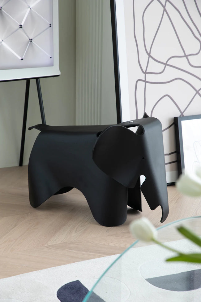 Black elephant-shaped stool in a room with a glass table and abstract wall art.