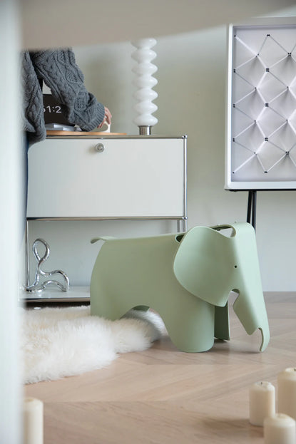 Green elephant plastic green stool toy, on a wooden floor with a white cabinet and decorative items in the background.