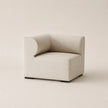 A modern low-profile accent chair upholstered in a light beige textured fabric, featuring a curved backrest and dark square feet, set against a solid warm beige background.