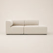A modern modular sectional sofa in a light beige textured fabric, shown from a front-facing three-quarter angle against a solid, warm off-white background.