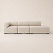A modern modular sectional sofa upholstered in a light beige textured fabric, shown against a matching minimalist beige background.