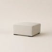 A square upholstered ottoman in a light beige fabric, shown from a three-quarter angle on a matching light beige studio background.