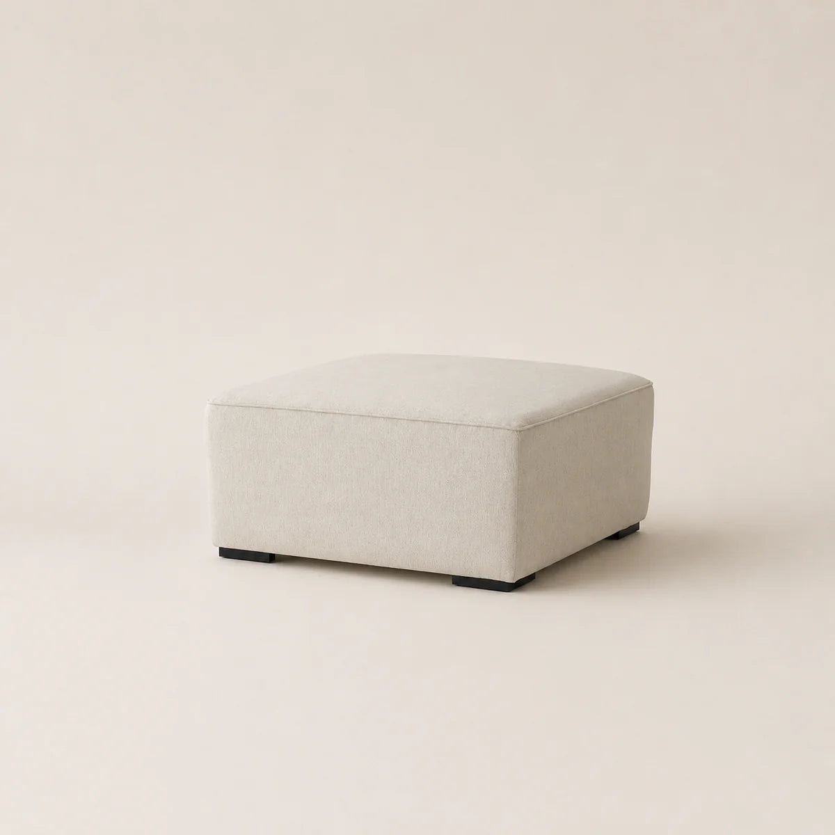 A square upholstered ottoman in a light beige fabric, shown from a three-quarter angle on a matching light beige studio background.
