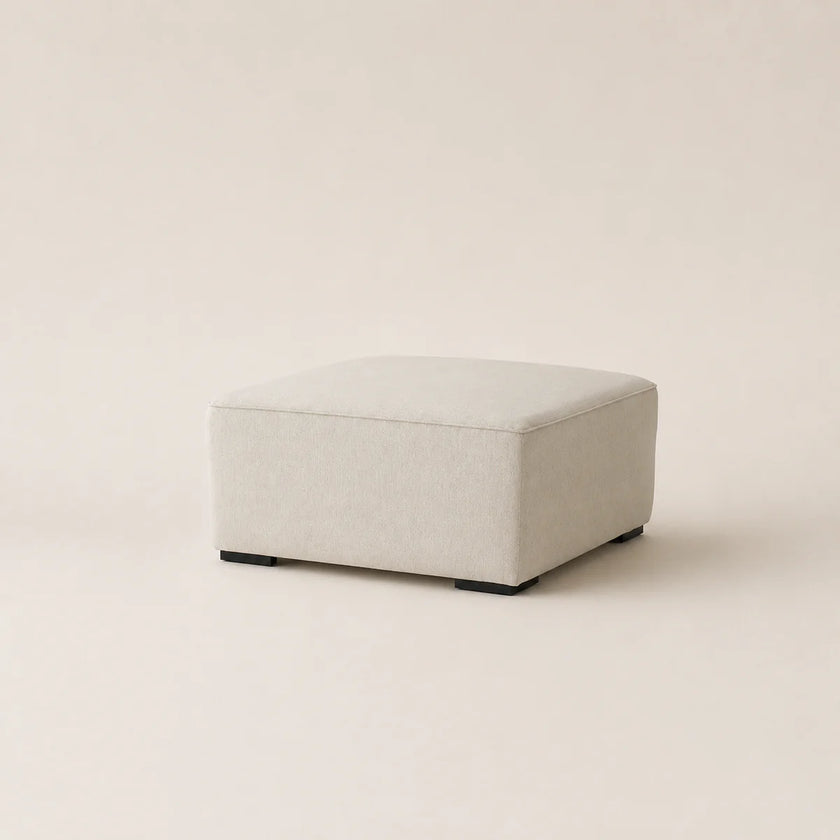 A square upholstered ottoman in a light beige fabric, shown from a three-quarter angle on a matching light beige studio background.