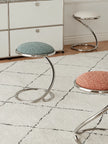 Three modern chrome stool stools with round textured fabric seats in blue-green, pink, and white, arranged on a white rug in a room setting.