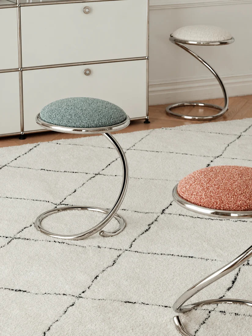 Three modern chrome stool stools with round textured fabric seats in blue-green, pink, and white, arranged on a white rug in a room setting.