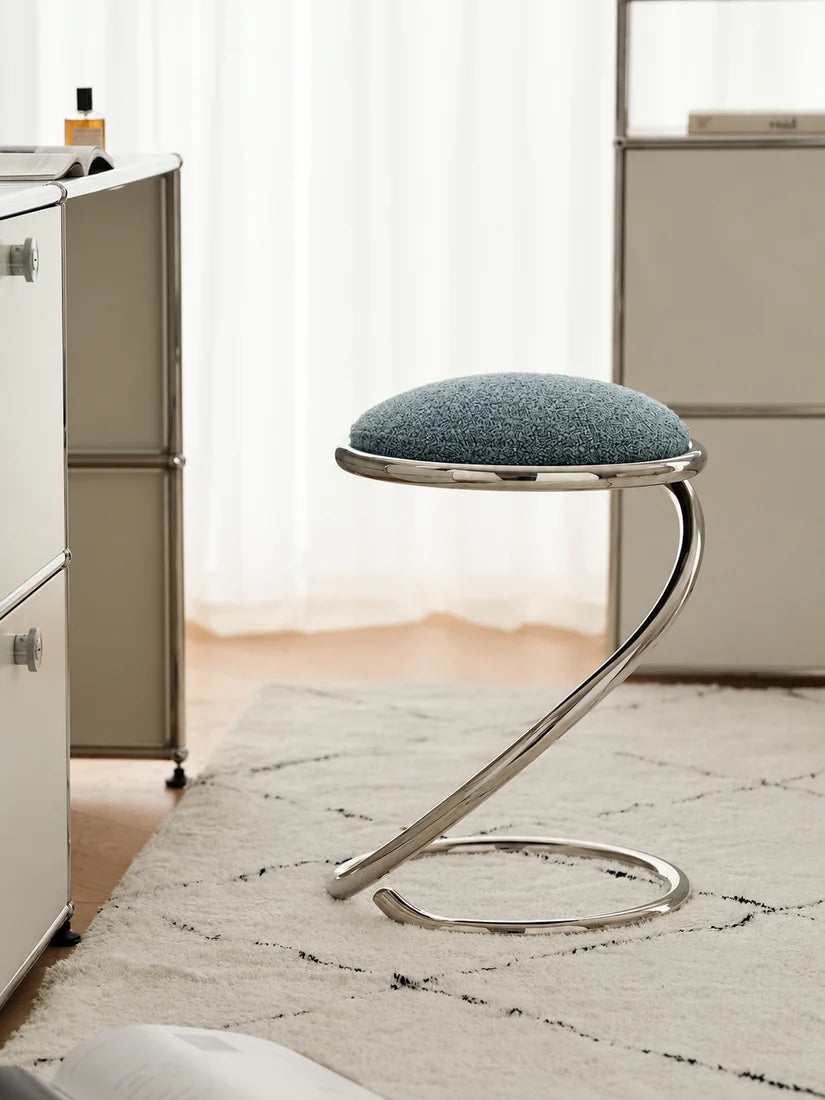 A modern curved chrome stool with a textured blue-gray upholstered seat cushion, shown in a bright, contemporary room setting with a white rug and light-colored furniture.