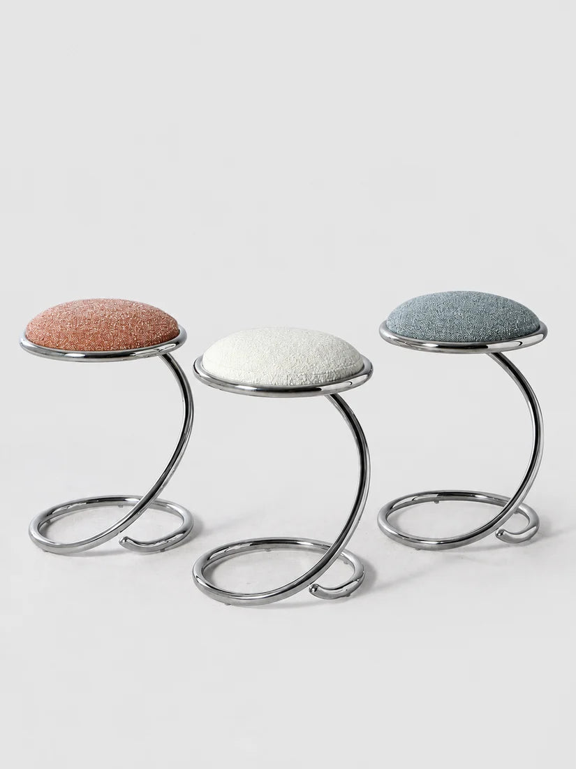 Three modern low stools with curved chrome metal bases and round upholstered seats in terracotta, off-white, and slate blue, arranged on a light gray studio background.