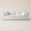 A modern low-profile white sofa with plush cushions and rolled arms, shown against a solid, warm off-white background.