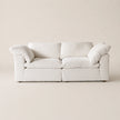 A modern two-seater sofa upholstered in a soft off-white fabric, featuring thick square back cushions, rounded armrests, and two matching throw pillows, set against a minimalist beige background.