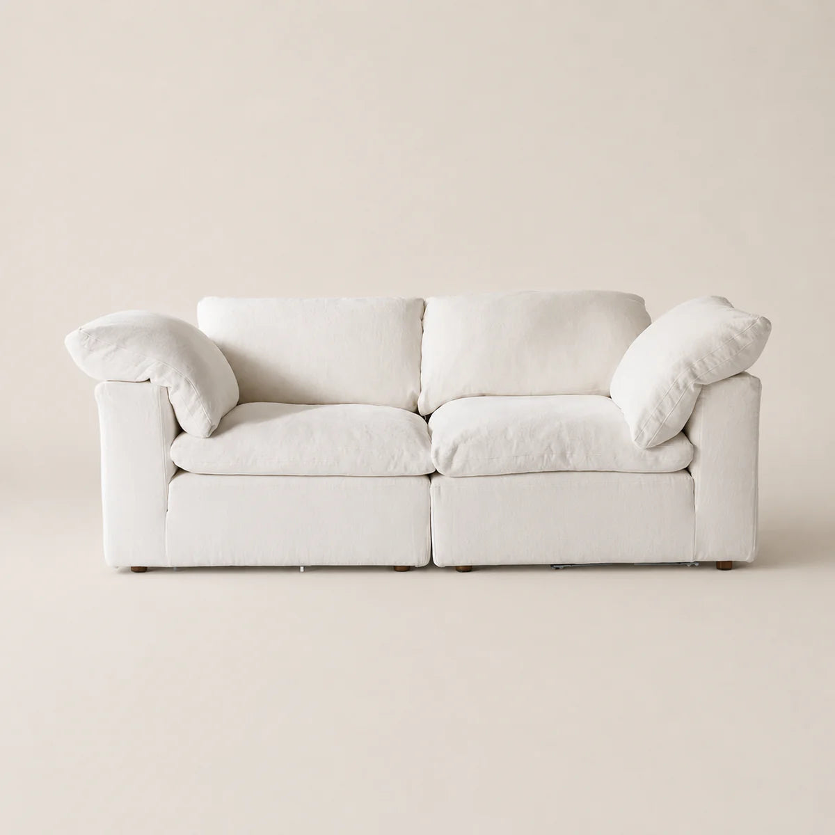 A modern two-seater sofa upholstered in a soft off-white fabric, featuring thick square back cushions, rounded armrests, and two matching throw pillows, set against a minimalist beige background.