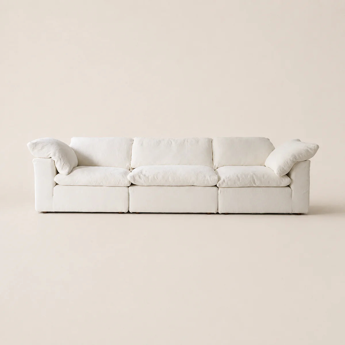 A modern three-seat sofa upholstered in off-white fabric, featuring plush cushions and rolled arms, set against a solid, warm off-white background.