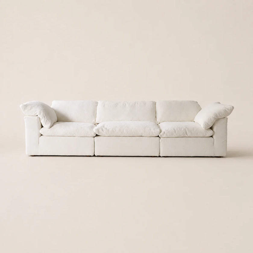 A modern three-seat sofa upholstered in off-white fabric, featuring plush cushions and rolled arms, set against a solid, warm off-white background.