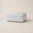 A square upholstered ottoman in a light white fabric, shown from a three-quarter angle on a matching light beige studio background.