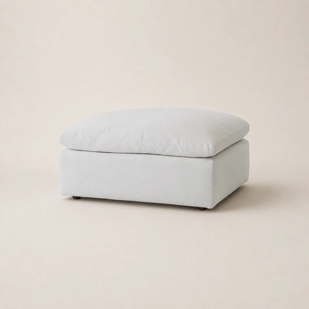A square upholstered ottoman in a light white fabric, shown from a three-quarter angle on a matching light beige studio background.