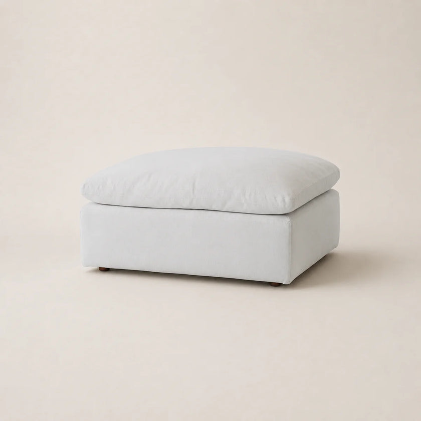 A square upholstered ottoman in a light white fabric, shown from a three-quarter angle on a matching light beige studio background.