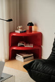 A bright red, two-tier modular side table styled in a minimalist room corner with a black floor lamp, a black leather chair, and books on its shelves.