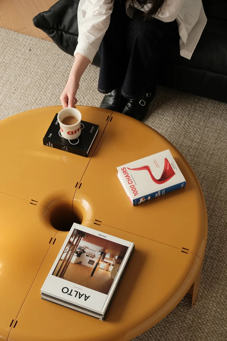 A round mustard-yellow coffee table in a modern living room setting, with a person's hand reaching for a cup on the table. Books are placed on the table, including an Alto magazine and a book with a red abstract cover.
