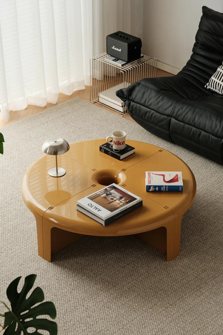 A round low coffee table in a warm honey-amber finish, styled in a modern living room setting with a black leather sofa, a Marshall speaker, and a textured beige rug.