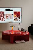 A glossy red round coffee table in a modern living room setting, featuring a metallic table lamp, eyeglasses, and books on its reflective surface.