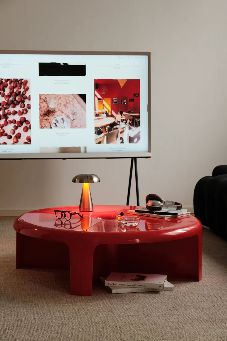 A glossy red round coffee table in a modern living room setting, featuring a metallic table lamp, eyeglasses, and books on its reflective surface.